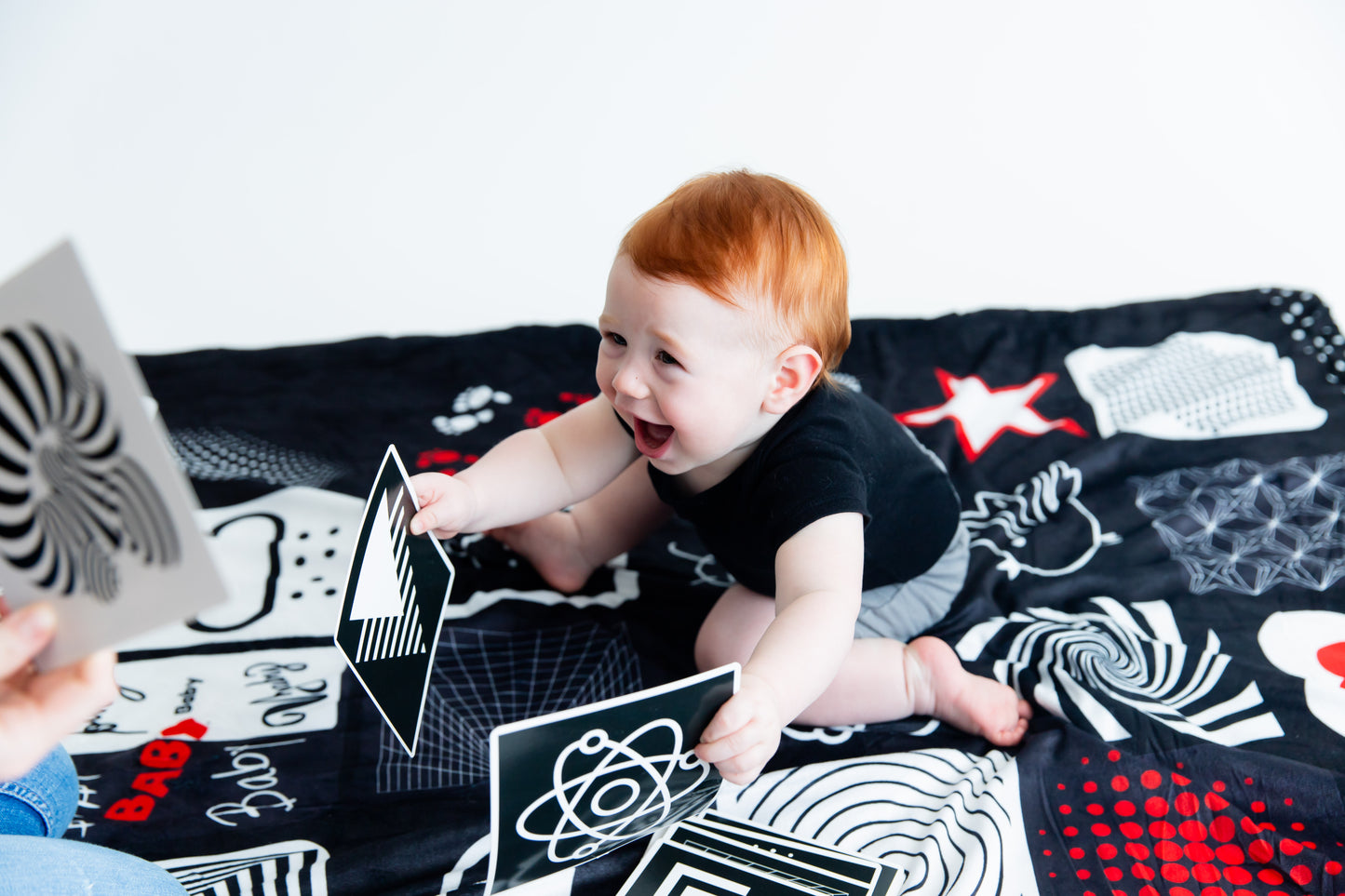 Mother showing Baby Geometrics Flashcards Set 1 to her adorable son, featuring high-contrast black-and-white designs crafted to enhance infant visual stimulation and foster parent-baby bonding moments. He is also sitting on the baby floor blanket. 