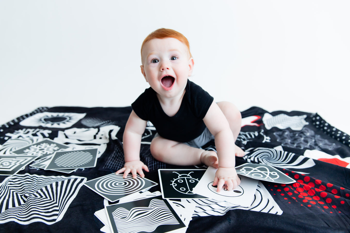 Redheaded baby smiling at the camera while sitting on the Baby Geometrics high-contrast floor blanket with flashcards, designed to enhance infant visual stimulation and cognitive growth.