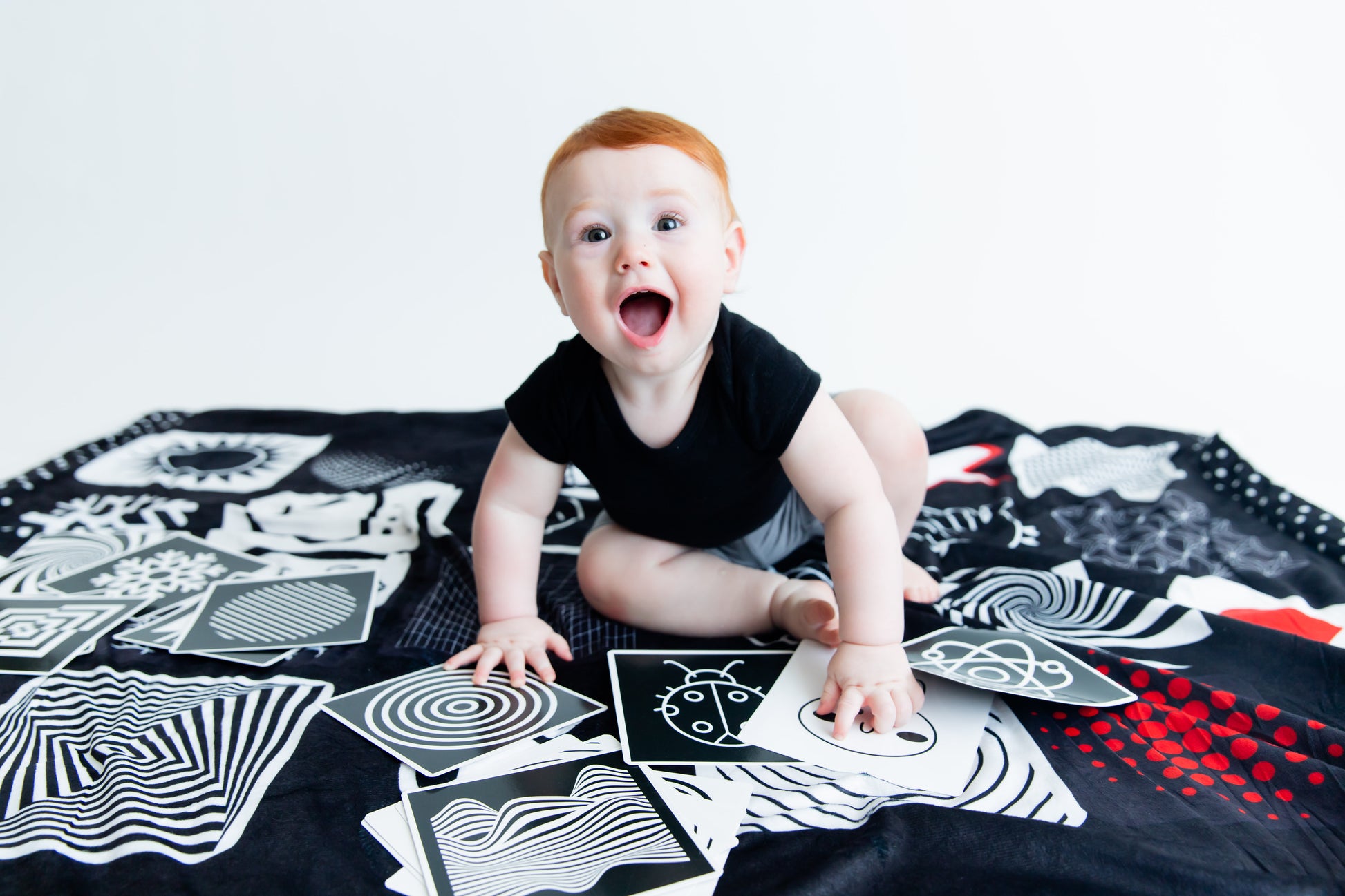 Redheaded baby smiling at the camera while sitting on the Baby Geometrics high-contrast floor blanket with flashcards, designed to enhance infant visual stimulation and cognitive growth.